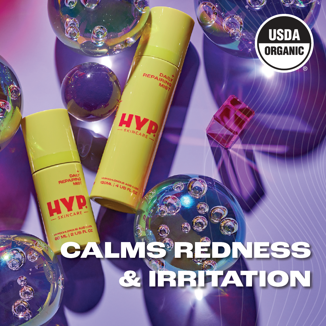 Two bottles of 'HYP Daily Repairing Mist' skincare product are displayed amongst reflective bubbles on a purplish surface with light flares. The text 'CALMS REDNESS & IRRITATION' in bold white letters is superimposed on the image, indicating the product's benefits. The 'USDA ORGANIC' seal is prominently featured on both the upper right corner and on the bottles, signifying the product's organic certification.