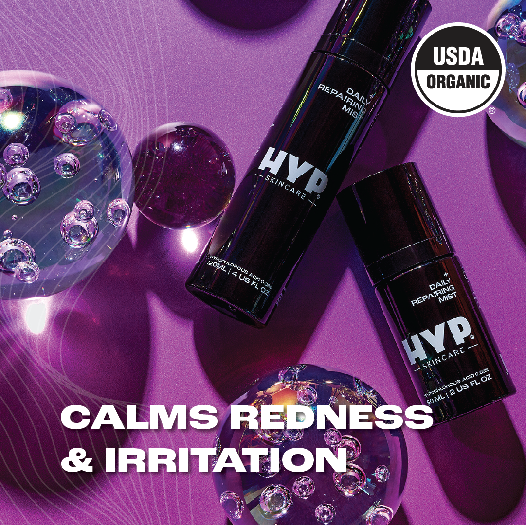 Two bottles of 'HYP Daily Repairing Mist' skincare product are displayed amongst reflective bubbles on a purplish surface with light flares. The text 'CALMS REDNESS & IRRITATION' in bold white letters is superimposed on the image, indicating the product's benefits. The 'USDA ORGANIC' seal is prominently featured on both the upper right corner and on the bottles, signifying the product's organic certification.