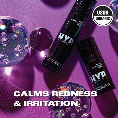 Two bottles of 'HYP Daily Repairing Mist' skincare product are displayed amongst reflective bubbles on a purplish surface with light flares. The text 'CALMS REDNESS & IRRITATION' in bold white letters is superimposed on the image, indicating the product's benefits. The 'USDA ORGANIC' seal is prominently featured on both the upper right corner and on the bottles, signifying the product's organic certification.