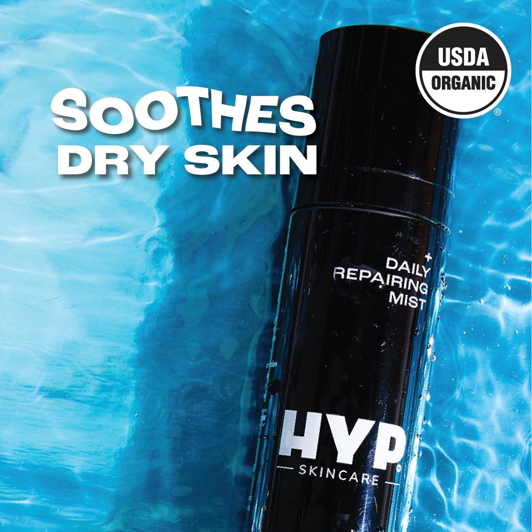 Close-up image of a black bottle of 'HYP Daily Repairing Mist' against a vivid blue, wavy background. Water droplets are visible on the bottle, suggesting hydration. Bold white text overlaid at the top reads 'SOOTHES DRY SKIN,' indicating the product's benefit. The 'USDA ORGANIC' seal is prominently displayed in the upper right corner, certifying the product's organic status.