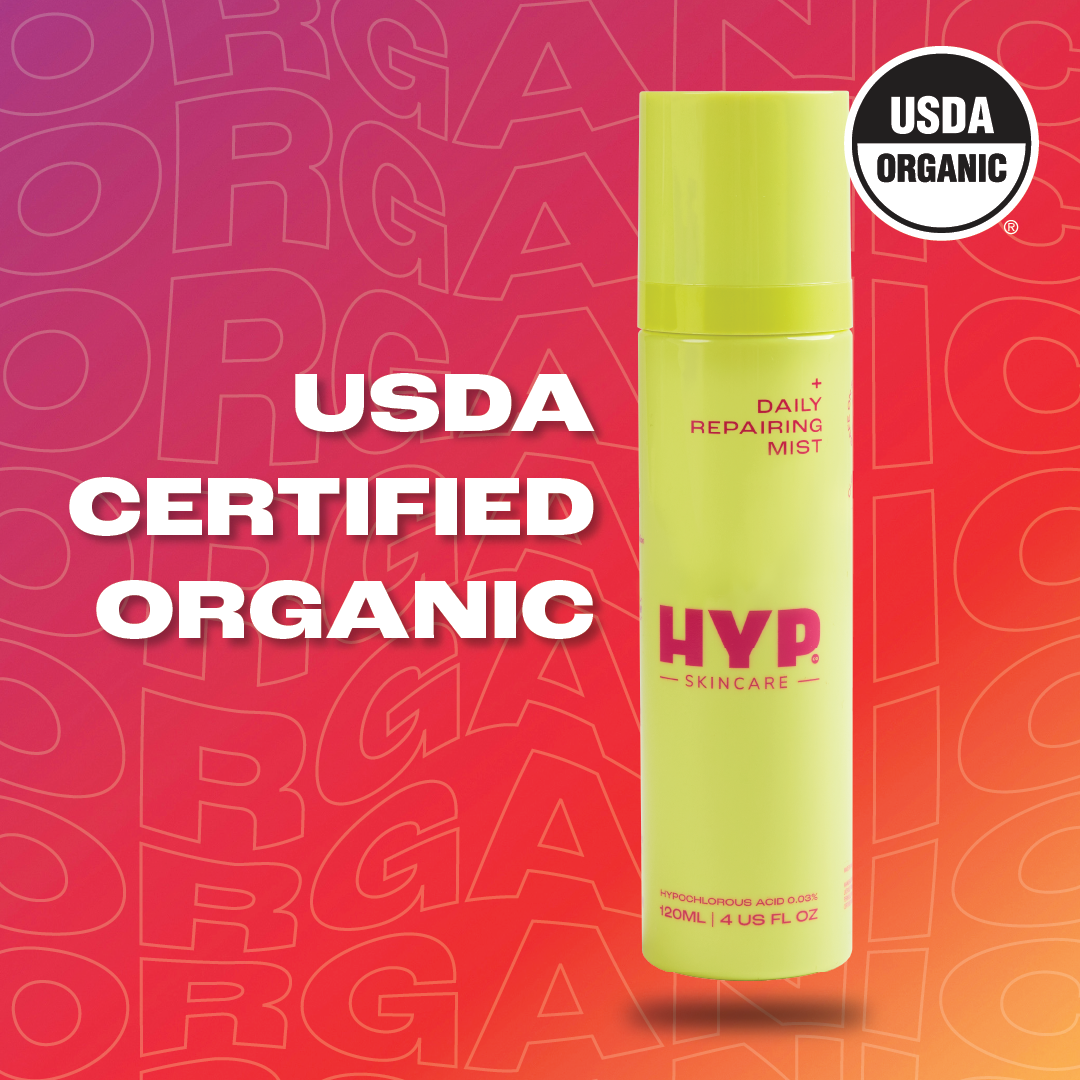 Image showcasing a yellow bottle of 'HYP Daily Repairing Mist' skincare product against a pink background patterned with multiple 'ORGANIC' texts. Prominent text overlay in white reads 'USDA CERTIFIED ORGANIC,' aligning with the 'USDA ORGANIC' seal displayed on both the upper right corner of the image and the bottle itself, highlighting the product's organic certification status.