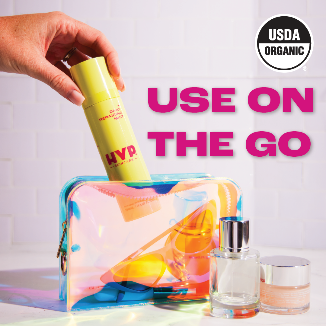 A hand is placing a yellow bottle of 'HYP Daily Repairing Mist' into a translucent, iridescent cosmetic bag alongside other skincare products. The image is set against a white backdrop with the magenta text 'USE ON THE GO' prominently displayed at the top, emphasizing the product's convenience for travel. The 'USDA ORGANIC' seal appears in the upper right corner, indicating the product's organic certification.