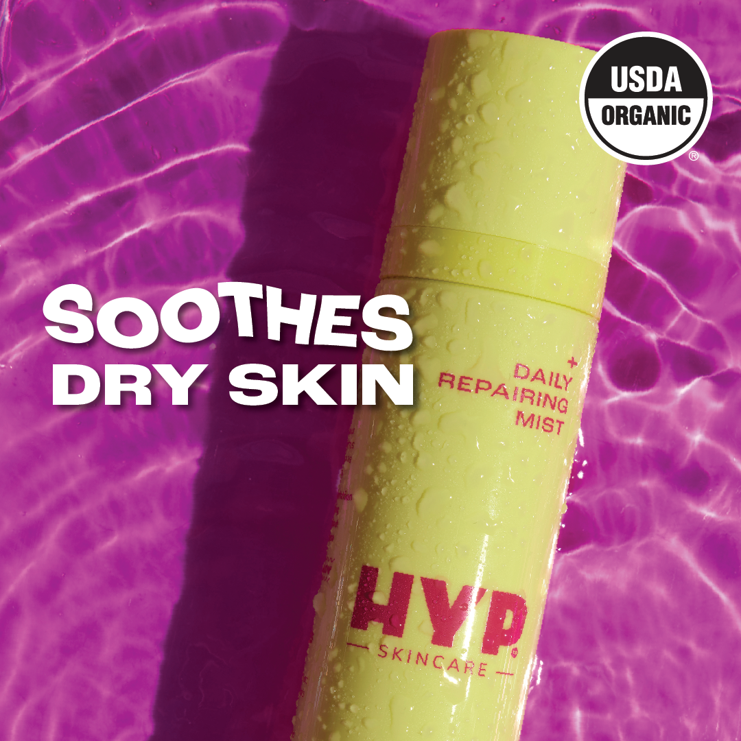 Close-up image of a yellow bottle of 'HYP Daily Repairing Mist' against a vivid purple, wavy background. Water droplets are visible on the bottle, suggesting hydration. Bold white text overlaid at the top reads 'SOOTHES DRY SKIN,' indicating the product's benefit. The 'USDA ORGANIC' seal is prominently displayed in the upper right corner, certifying the product's organic status